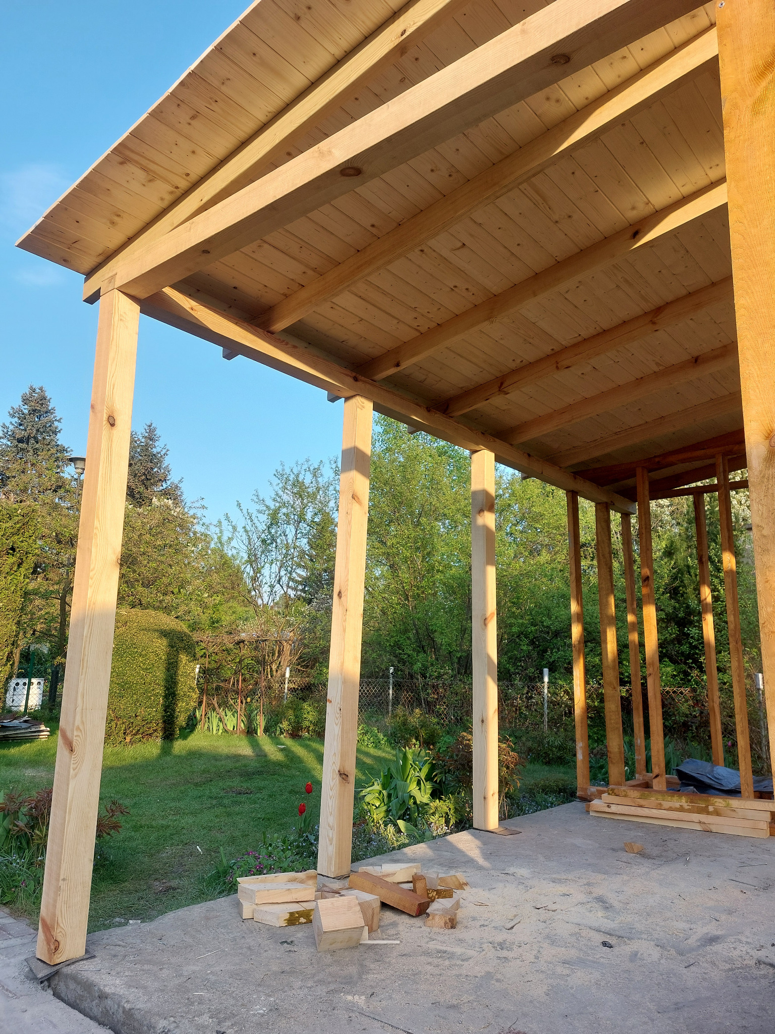 Wznoszenie drewnianej konstrukcji altany z widocznym zadaszeniem i słupami nośnymi na tle zielonego ogrodu w słoneczny dzień. Widoczne elementy konstrukcyjne i deski.
