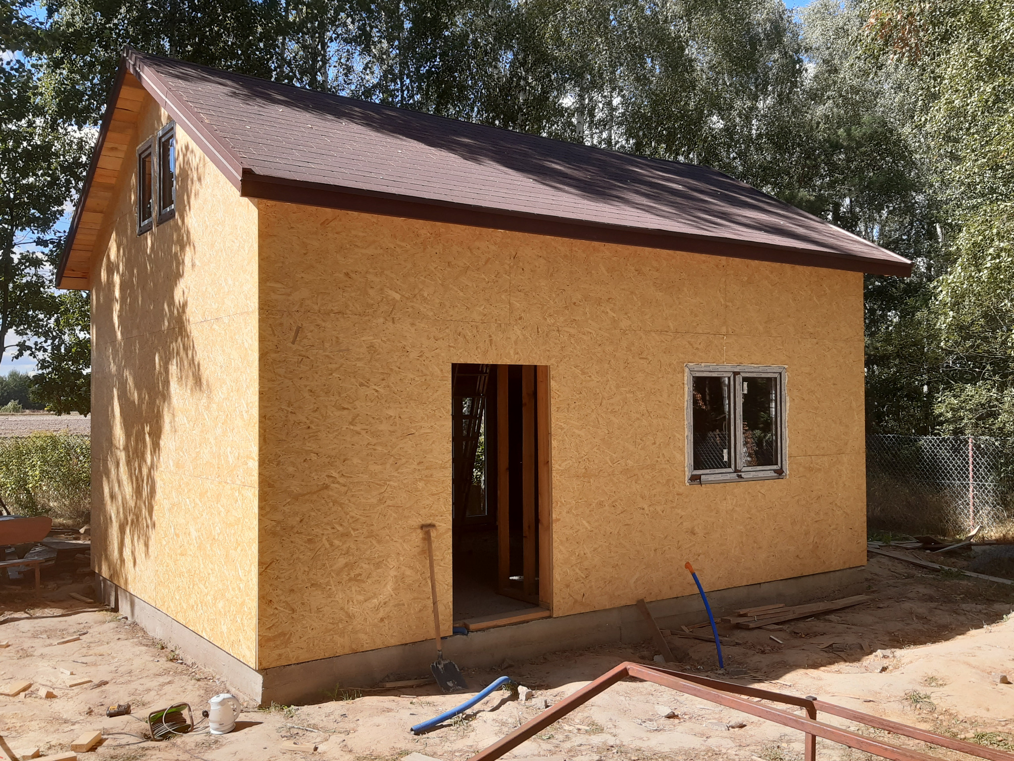 Surowy stan domu szkieletowego z brązowym dachem, widoczne płyty OSB, okno i otwór drzwiowy. Na ziemi narzędzia i elementy konstrukcyjne. Tło: drzewa.