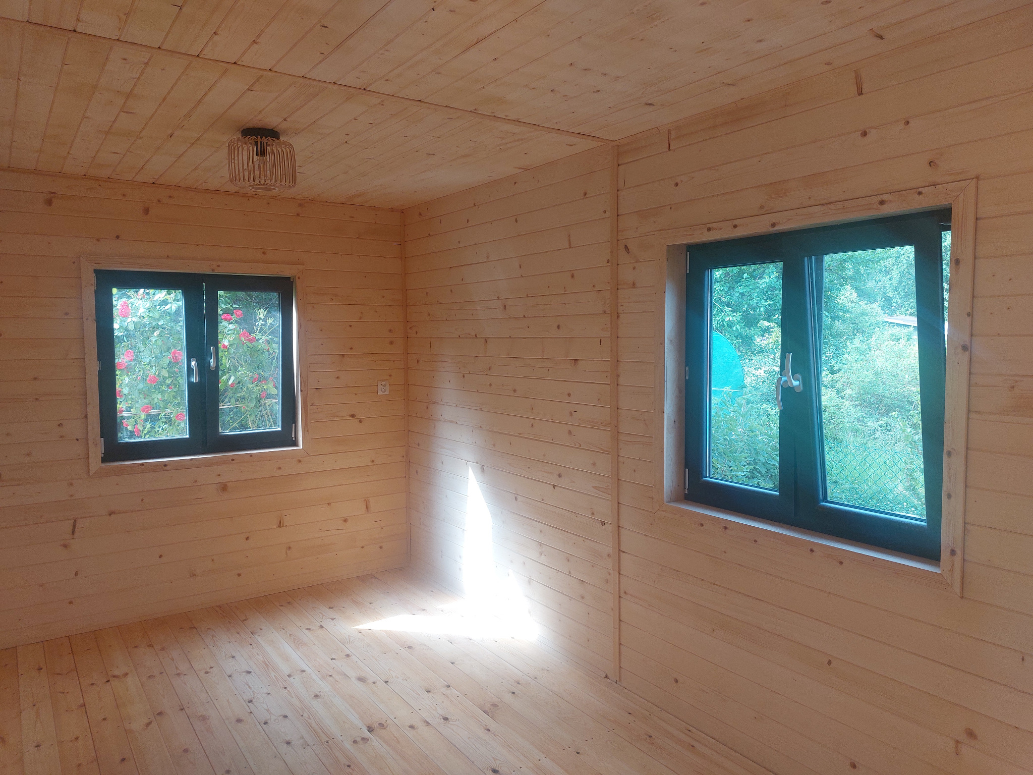 Wnętrze drewnianego domku z dwoma oknami, przez które widać zieleń. Ściany, podłoga i sufit wyłożone jasnym drewnem. Na suficie widoczna lampa z abażurem.