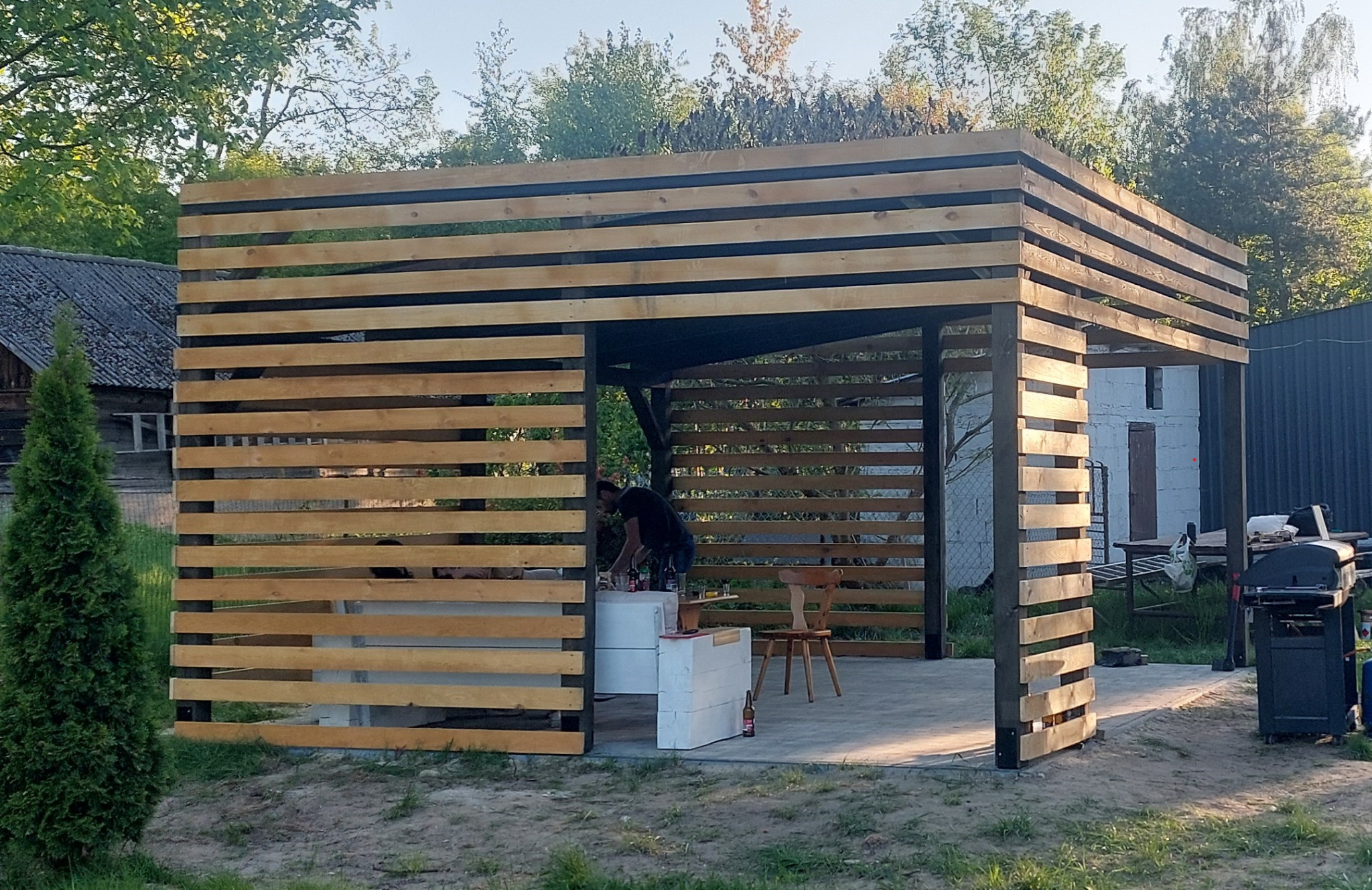 Nowoczesna altana z drewnianymi lamelami, częściowo zasłaniająca osobę przygotowującą napoje. Widoczny grill i elementy wyposażenia ogrodu. Ujęcie w słoneczny dzień.