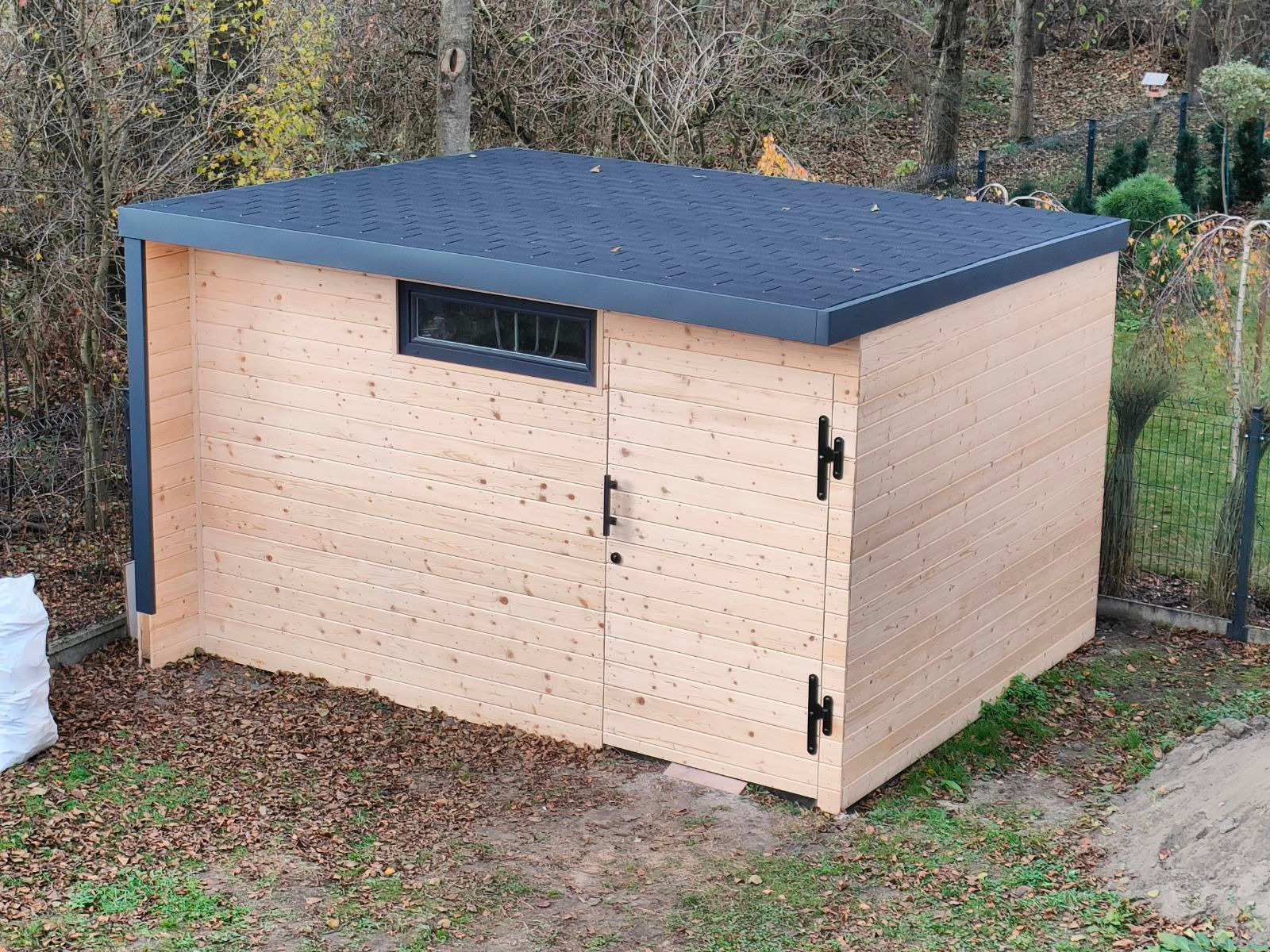 Nowoczesna, drewniana altana ogrodowa z płaskim dachem i oknem, wykończona w minimalistycznym stylu, otoczona jesiennym krajobrazem.