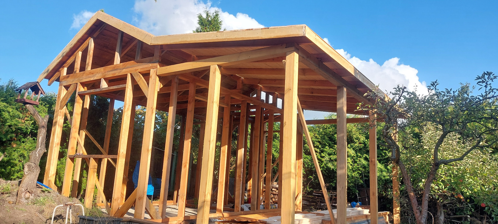Drewniana konstrukcja altany w ogrodzie, widoczne słupy i zadaszenie w trakcie budowy, na tle zieleni i błękitnego nieba z chmurami.