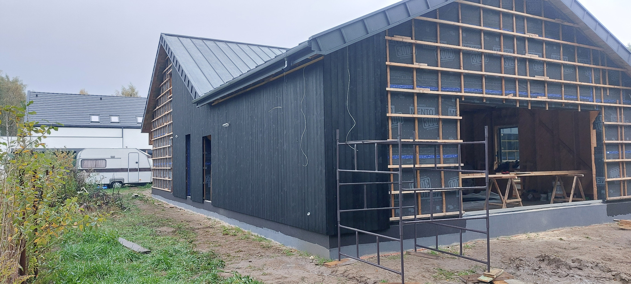 Budowa domu szkieletowego z czarną elewacją i widoczną konstrukcją drewnianą. Na pierwszym planie rusztowanie, w tle dom i przyczepa kempingowa.