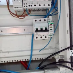 Rozdzielnica elektryczna z zamontowanymi bezpiecznikami, wyłącznikiem różnicowoprądowym i przewodami w kolorach niebieskim, brązowym i czarnym, zamontowana w białej obudowie.