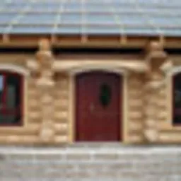 Elewacja budynku z jasnego kamienia z ciemnoczerwonymi, drewnianymi drzwiami wejściowymi i dwoma symetrycznie umieszczonymi, wąskimi oknami po bokach. Nad drzwiami i oknami widoczne drewniane...