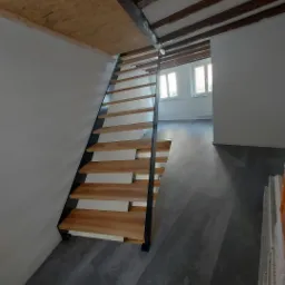 Nowoczesne schody z drewnianymi stopniami i czarną metalową konstrukcją boczną, zamontowane w jasnym wnętrzu z drewnianym stropem i szarą podłogą.