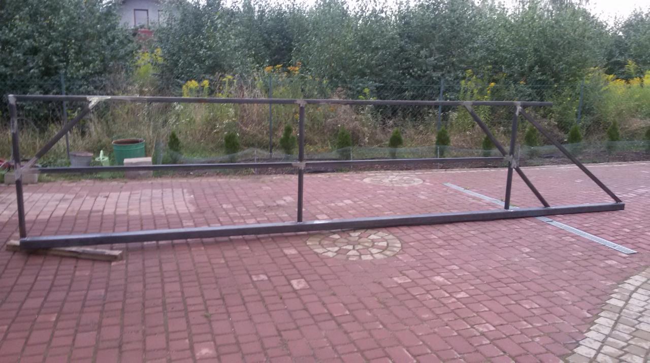 Surowa, spawana konstrukcja metalowa ogrodzenia leżąca na czerwonej kostce brukowej, z widocznymi śladami spawania i elementami podtrzymującymi, w tle zielony ogród i dom.