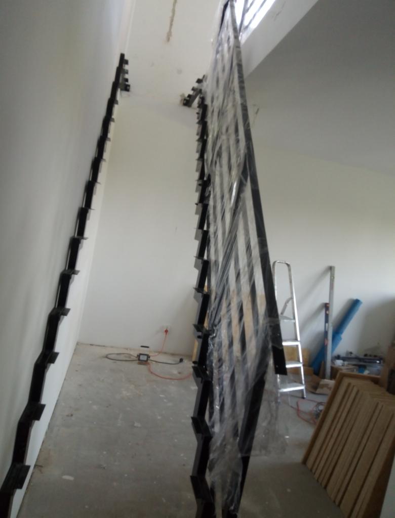 Montaż czarnej, metalowej konstrukcji schodów z elementami mocującymi do ściany, zabezpieczonej folią, w surowym wnętrzu z drabiną w tle.