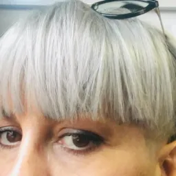 Zbliżenie na twarz kobiety z krótką, srebrną fryzurą typu bowl cut i okularami przeciwsłonecznymi na głowie, widoczne brązowe oczy i delikatny makijaż.