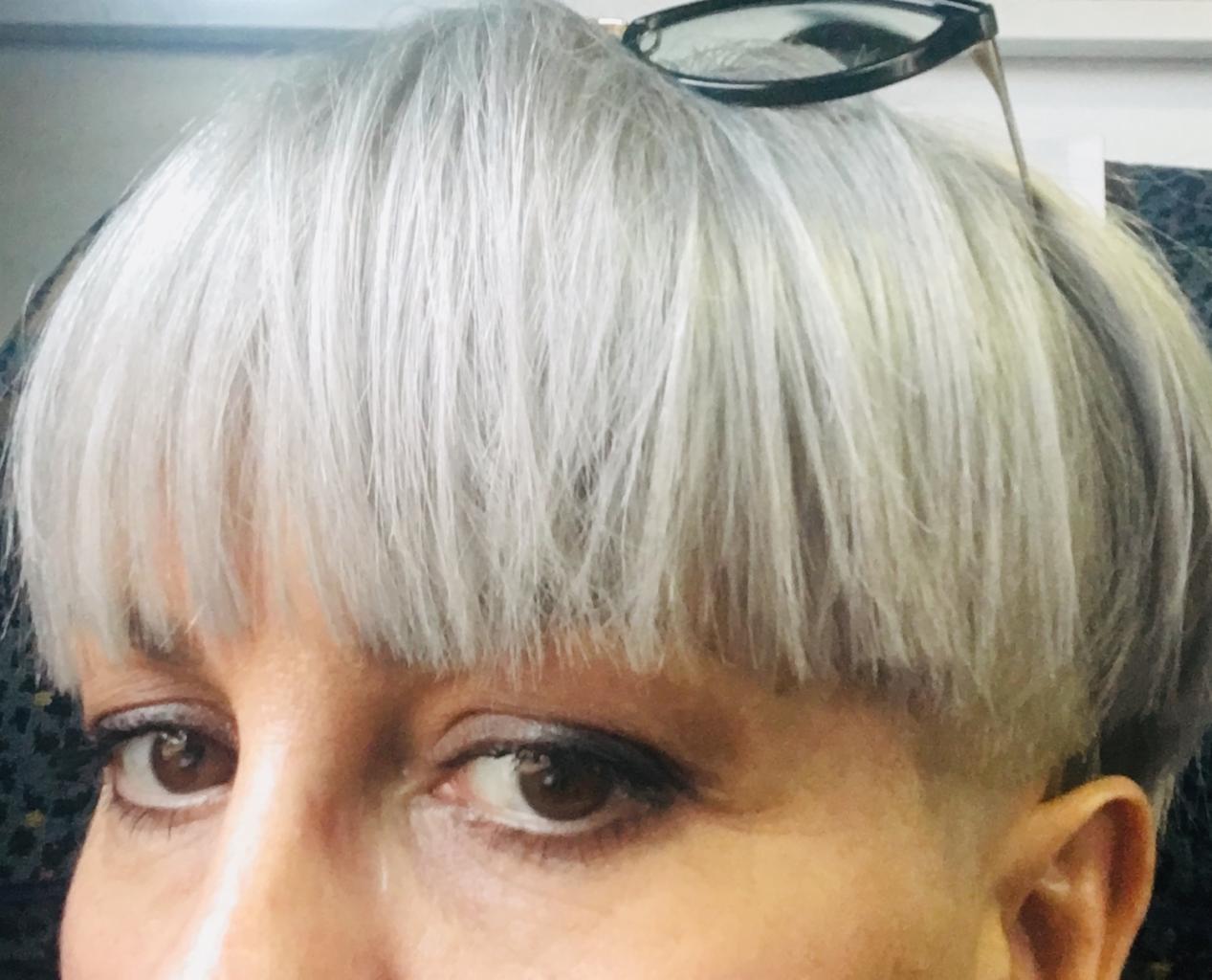 Zbliżenie na twarz kobiety z krótką, srebrną fryzurą typu bowl cut i okularami przeciwsłonecznymi na głowie, widoczne brązowe oczy i delikatny makijaż.