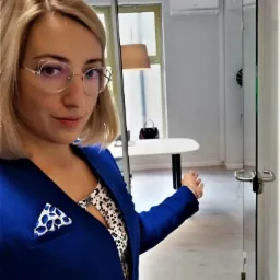 Kobieta w okularach i niebieskiej marynarce stoi w biurze, otwierając szklane drzwi. W tle białe biurko i klimatyzator.