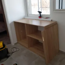 Montaż szafki z drewnopodobnej płyty pod oknem w Trzebiatowie, widoczny wkrętak akumulatorowy na parapecie i odkurzacz Stanley na podłodze.