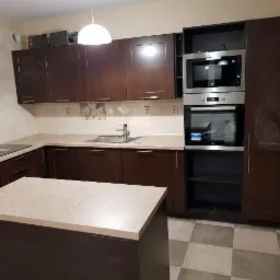 Ciemnobrązowa zabudowa kuchenna z wyspą, beżowy blat, płyta indukcyjna i piekarnik w zabudowie, na ścianie płytki z motywem kwiatowym, podłoga w szachownicę.