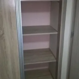 Wnętrze szafy przesuwnej z półkami o jasnym odcieniu drewna, widoczne prowadnice drzwi i różowe tło ścian wewnątrz szafy.