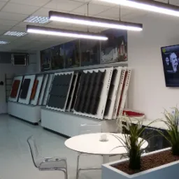 Ekspozycja różnych modeli pokryć dachowych w salonie firmowym w Żyrardowie. W tle widoczne zdjęcia domów z różnymi rodzajami dachów, a na ścianie telewizor z aktualną listą odtwarzania.