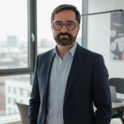 Portret mężczyzny w okularach i garniturze na tle okna biurowego. Profesjonalny wygląd, sugerujący doradztwo biznesowe lub finansowe.