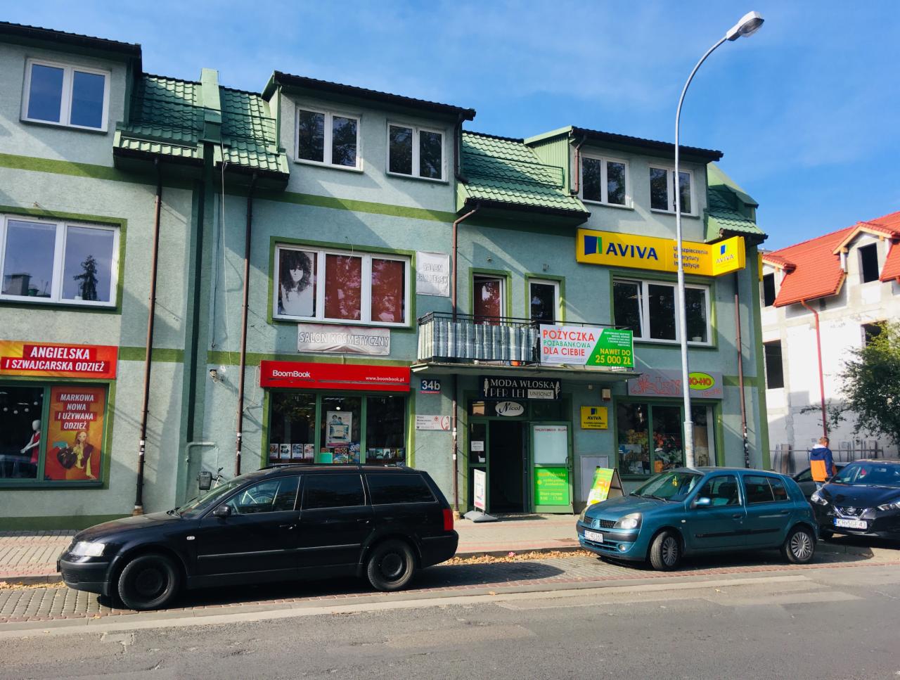 Budynek usługowy z szyldami różnych firm, w tym reklama pożyczki pozabankowej do 25 000 zł oraz salonu kosmetycznego, z zaparkowanymi samochodami na pierwszym planie.