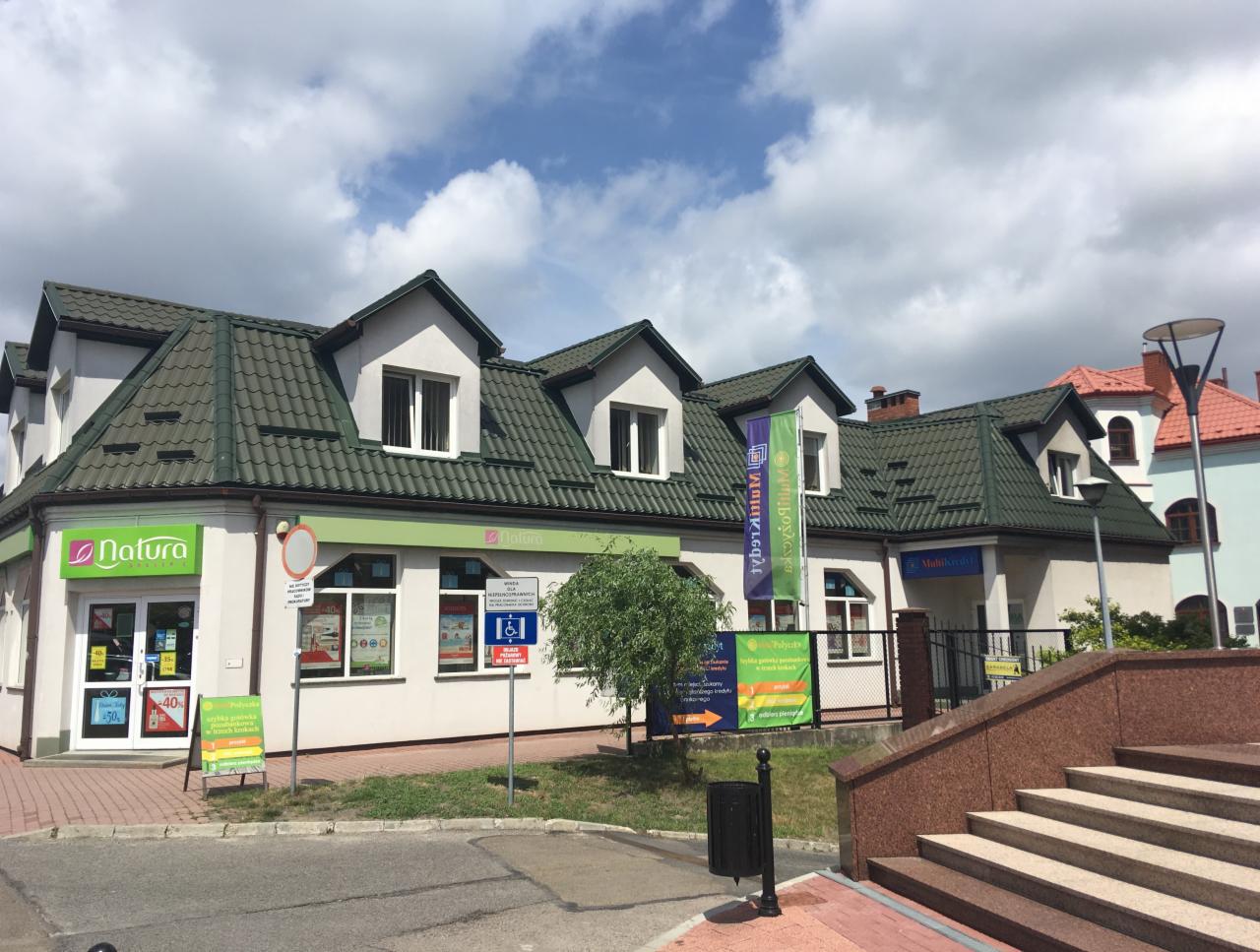 Fasada budynku z szyldami 'Natura' i 'MultiKredyt', zielony dach, białe ściany, flaga reklamowa z napisem 'MultiKredyt', małe drzewo przed budynkiem, schody prowadzące do wejścia.