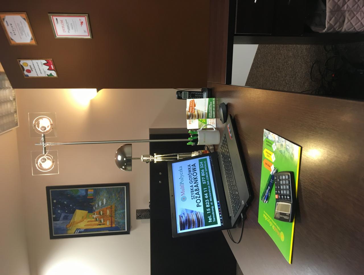 Biuro pożyczkowe: Laptop z wyświetloną ofertą, ulotka z kalkulatorem, lampa biurkowa, dyplomy na ścianie i przybory biurowe na drewnianym biurku.