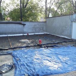 SzymonT - Przygotowanie podłoża pod wylewkę betonową: siatka zbrojeniowa na warstwie izolacyjnej, widoczna studzienka kanalizacyjna i niebieska plandeka zabezpieczająca kostkę brukową.