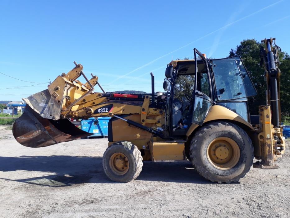 Żółta koparko-ładowarka Caterpillar 432E z otwartą kabiną i łyżką z przodu, stojąca na ziemi z widocznymi śladami użytkowania, na tle błękitnego nieba i kontenerów.