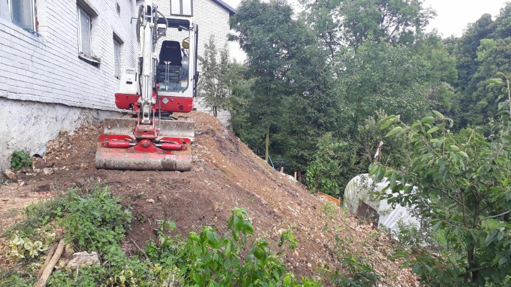 Czerwono-biała mini koparka na nierównym terenie przy fundamencie budynku, przygotowująca teren pod dalsze prace.