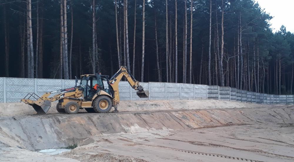 Żółta koparko-ładowarka CAT 432E wyrównuje teren przy betonowym ogrodzeniu panelowym na tle wysokiego lasu sosnowego.