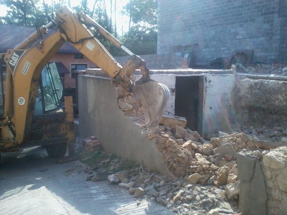 Żółta koparko-ładowarka CAT kruszy betonową ścianę, tworząc stertę gruzu przed wejściem do piwnicy budynku z cegły.
