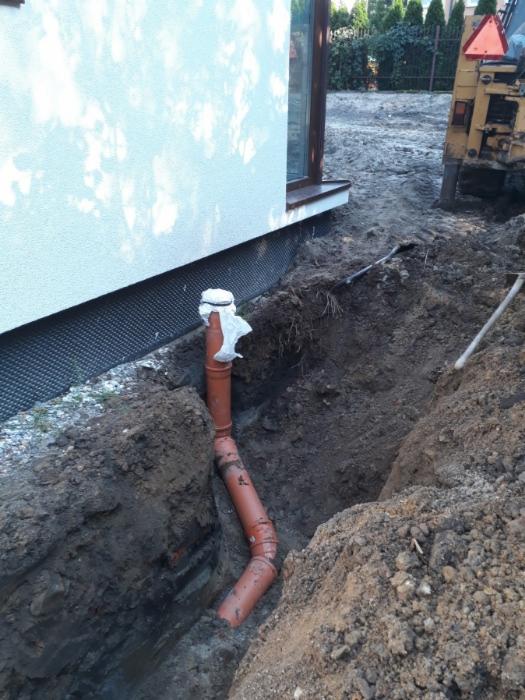 Pomarańczowa rura kanalizacyjna ułożona w wykopie obok budynku z ociepleniem fundamentów, widoczny fragment koparki w tle.