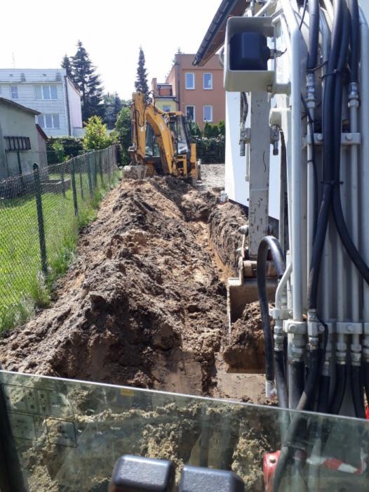 Żółta koparka głębiąca wąski wykop w ziemi, widok z kabiny innej maszyny, ogrodzenie z siatki i budynki w tle.