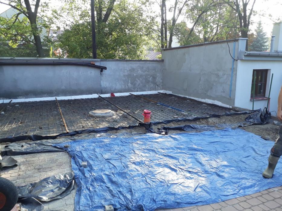 Przygotowanie podłoża pod wylewkę betonową: siatka zbrojeniowa na warstwie izolacyjnej, widoczna studzienka kanalizacyjna i niebieska plandeka zabezpieczająca kostkę brukową.