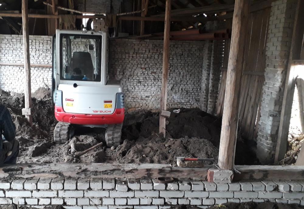 Wnętrze starego budynku w trakcie wyburzania, z widoczną mini-koparką Takeuchi na tle ceglanej ściany i sterty gruzu.