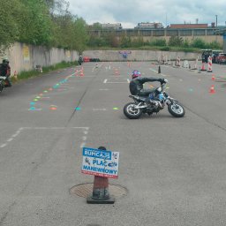 Nauka Jazdy RUMCAJS Marcin Baranowski - Kursant na małym motocyklu ćwiczy manewry na placu, otoczonym pachołkami. W tle budynki i drzewa. Tablica informacyjna szkoły jazdy na pierwszym planie.