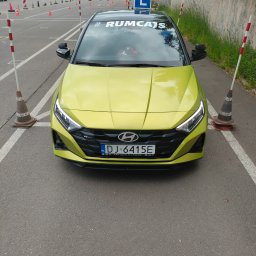 Nauka Jazdy RUMCAJS Marcin Baranowski - Żółty Hyundai i20 z naklejką #RUMCATS na szybie, stojący na placu manewrowym szkoły jazdy, otoczony pachołkami i słupkami. Widoczna tablica z literą 'L'.