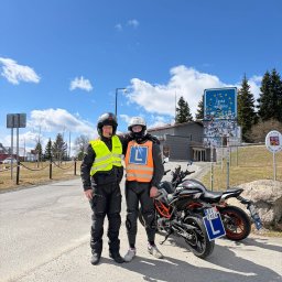 Nauka Jazdy RUMCAJS Marcin Baranowski - Instruktor i kursant szkoły jazdy motocyklem, obaj w kaskach i kamizelkach odblaskowych, stojący obok motocykla z tablicą 'L' na tle znaku granicznego.