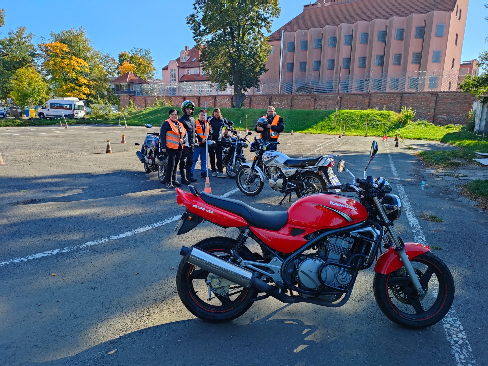 Czerwony motocykl Kawasaki ER-5 na placu manewrowym, grupa osób w kamizelkach odblaskowych i kaskach, w tle budynek z czerwonej cegły