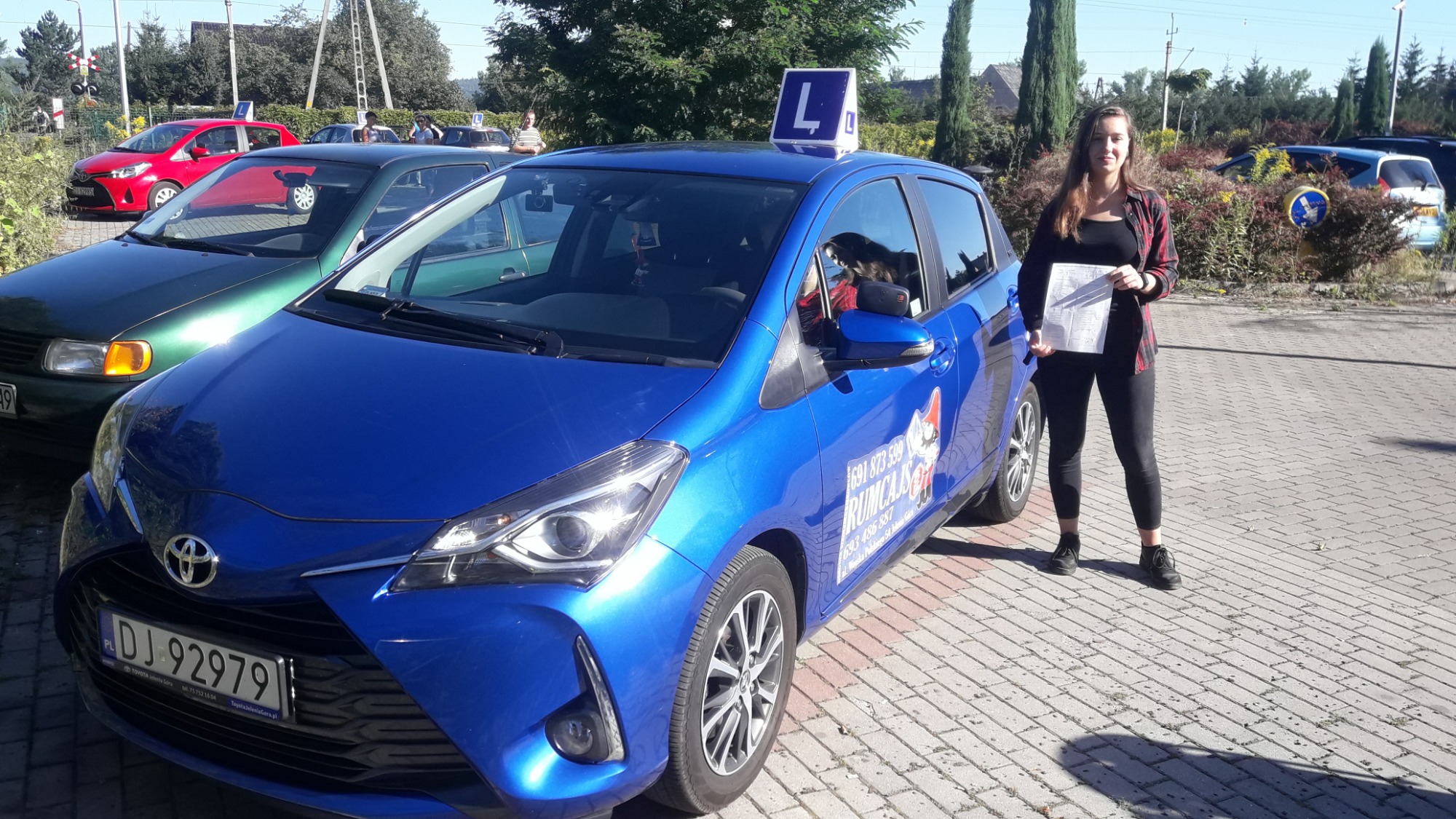 Niebieski samochód Toyota Yaris z literą 'L' na dachu, zaparkowany na brukowanym placu, obok stoi uśmiechnięta kobieta trzymająca dokumenty, w tle inne pojazdy i drzewa.