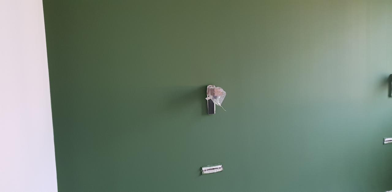 Świeżo pomalowana na oliwkowo-zielony kolor ściana z zabezpieczonymi folią gniazdkami elektrycznymi i włącznikami, widoczny fragment białej ściany z lewej strony.