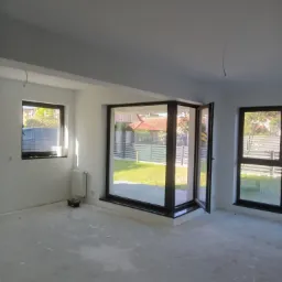 Jasne, przestronne wnętrze po remoncie. Białe ściany, duże okna z czarnymi ramami, surowa podłoga. Widok na ogród przez otwarte drzwi balkonowe.