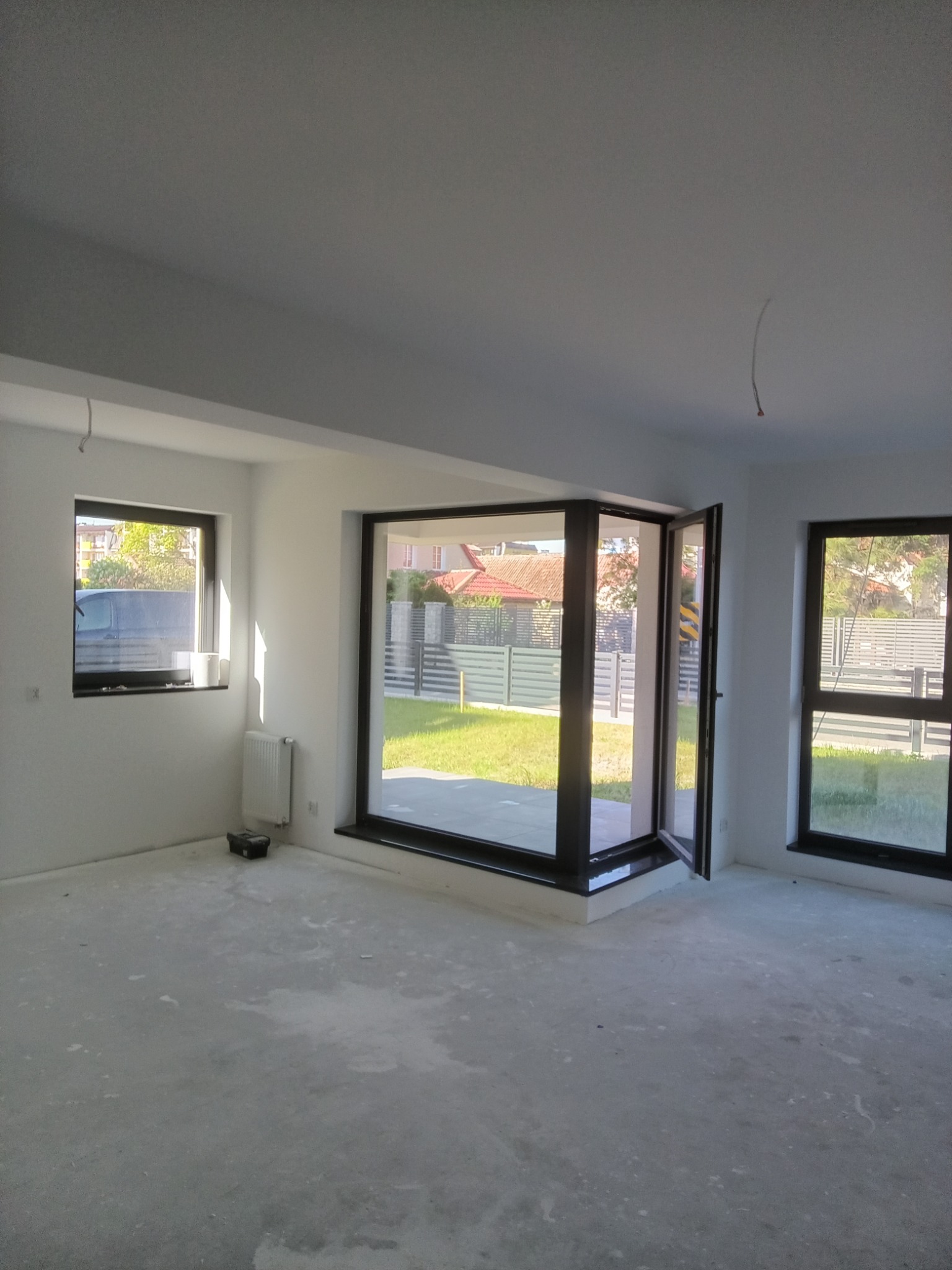 Jasne, przestronne wnętrze po remoncie. Białe ściany, duże okna z czarnymi ramami, surowa podłoga. Widok na ogród przez otwarte drzwi balkonowe.