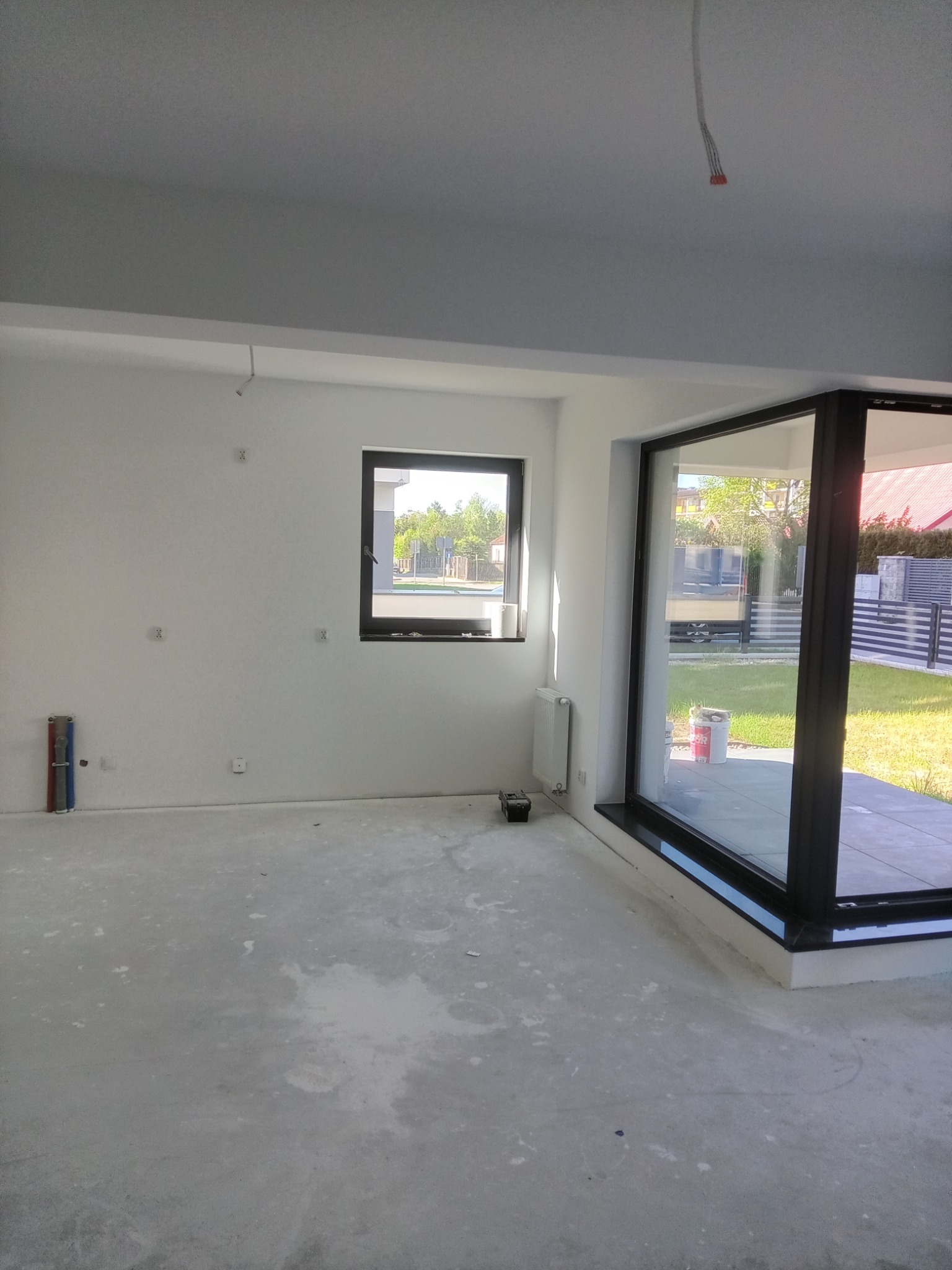 Wnętrze pokoju w stanie deweloperskim z oknem i drzwiami balkonowymi. Ściany białe, surowe. Na podłodze widoczne ślady po pracach budowlanych. Grzejnik pod oknem.