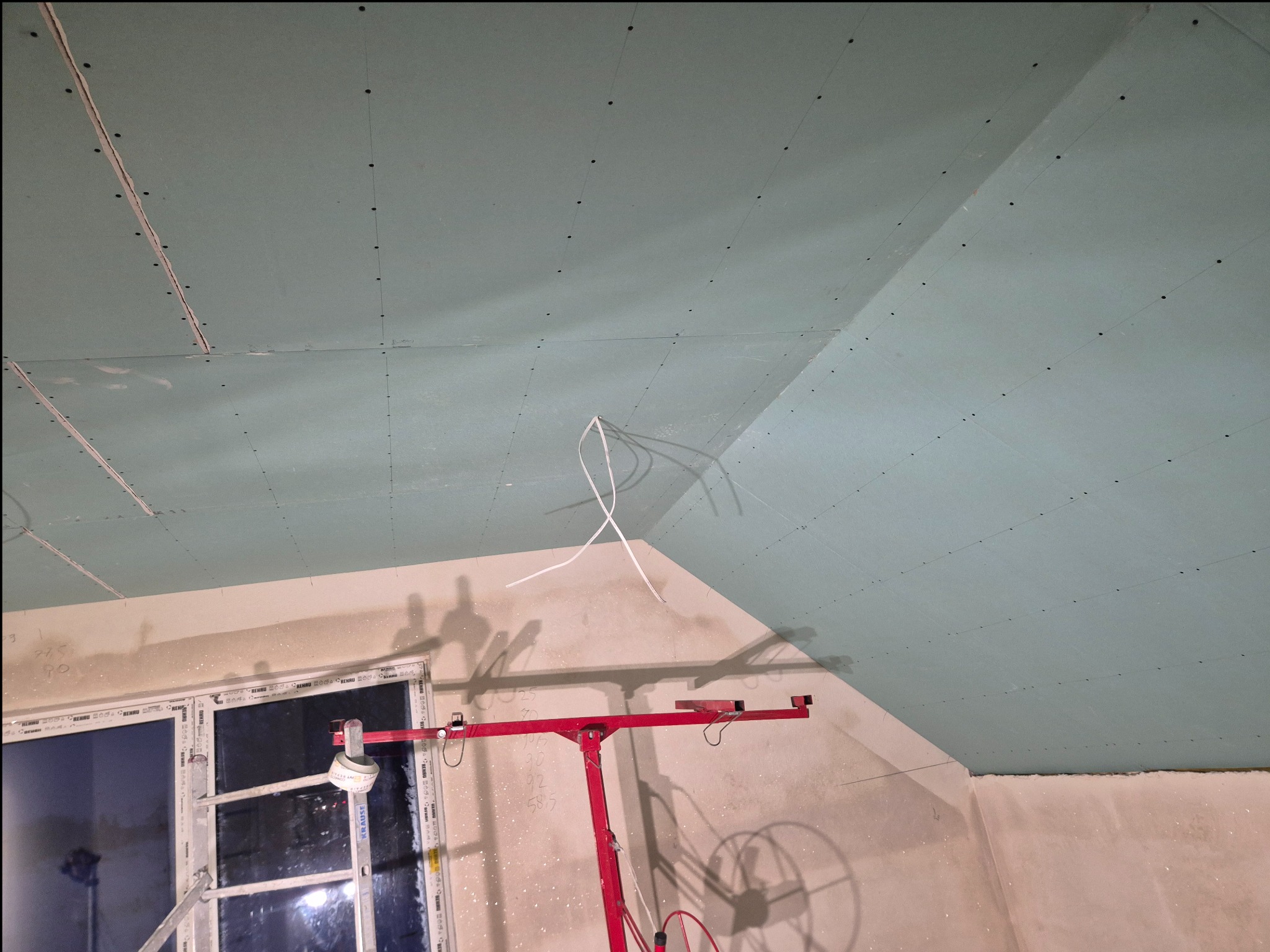 Montaż płyt karton-gips na suficie poddasza, widoczne łączenia i wkręty, czerwony podnośnik do płyt. Ściany przygotowane do dalszych prac wykończeniowych.