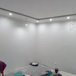 Pomalowane na biało ściany pokoju z zamontowanym oświetleniem LED, podłoga zabezpieczona folią malarską, widoczne akcesoria malarskie.