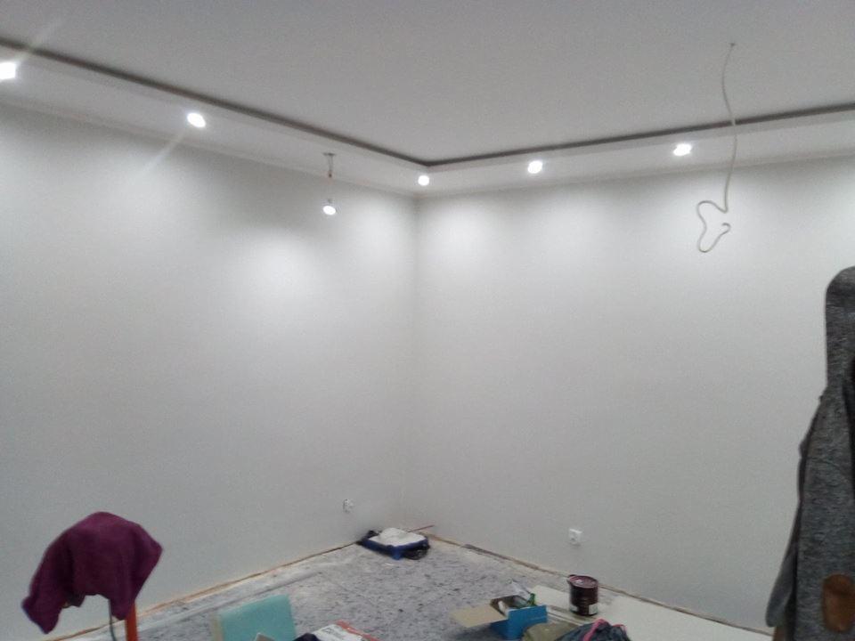 Pomalowane na biało ściany pokoju z zamontowanym oświetleniem LED, podłoga zabezpieczona folią malarską, widoczne akcesoria malarskie.