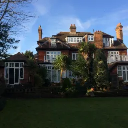 Duży, dwupiętrowy dom z czerwonej cegły z białymi oknami, balkonami i palmami w ogrodzie, widziany z poziomu trawnika w słoneczny dzień.