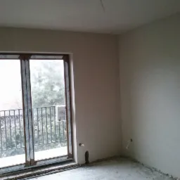 Wnętrze pokoju po tynkowaniu maszynowym; widoczne gładkie ściany, okno balkonowe z widokiem na zewnątrz, oraz wystające kable elektryczne.