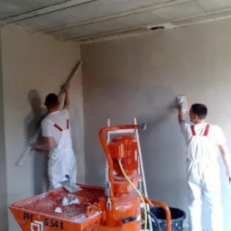 Dwóch pracowników w białych kombinezonach wygładza ściany wewnątrz budynku za pomocą maszyny do nakładania tynku i narzędzi ręcznych.