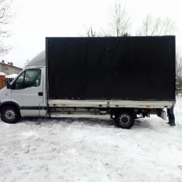 Biały bus z czarną plandeką zaparkowany na śniegu, widoczne nogi osoby stojącej za pojazdem.