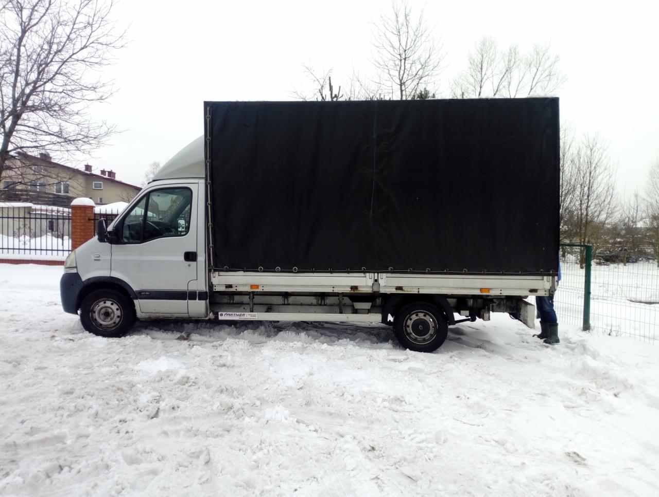 Srebrny bus dostawczy z czarną plandeką zaparkowany na śniegu w zimowy dzień, widoczne fragmenty ogrodzenia i zabudowań w tle.