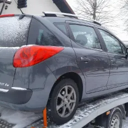 Szary samochód Peugeot 207 SW pokryty śniegiem, transportowany na lawecie w zimowy dzień.